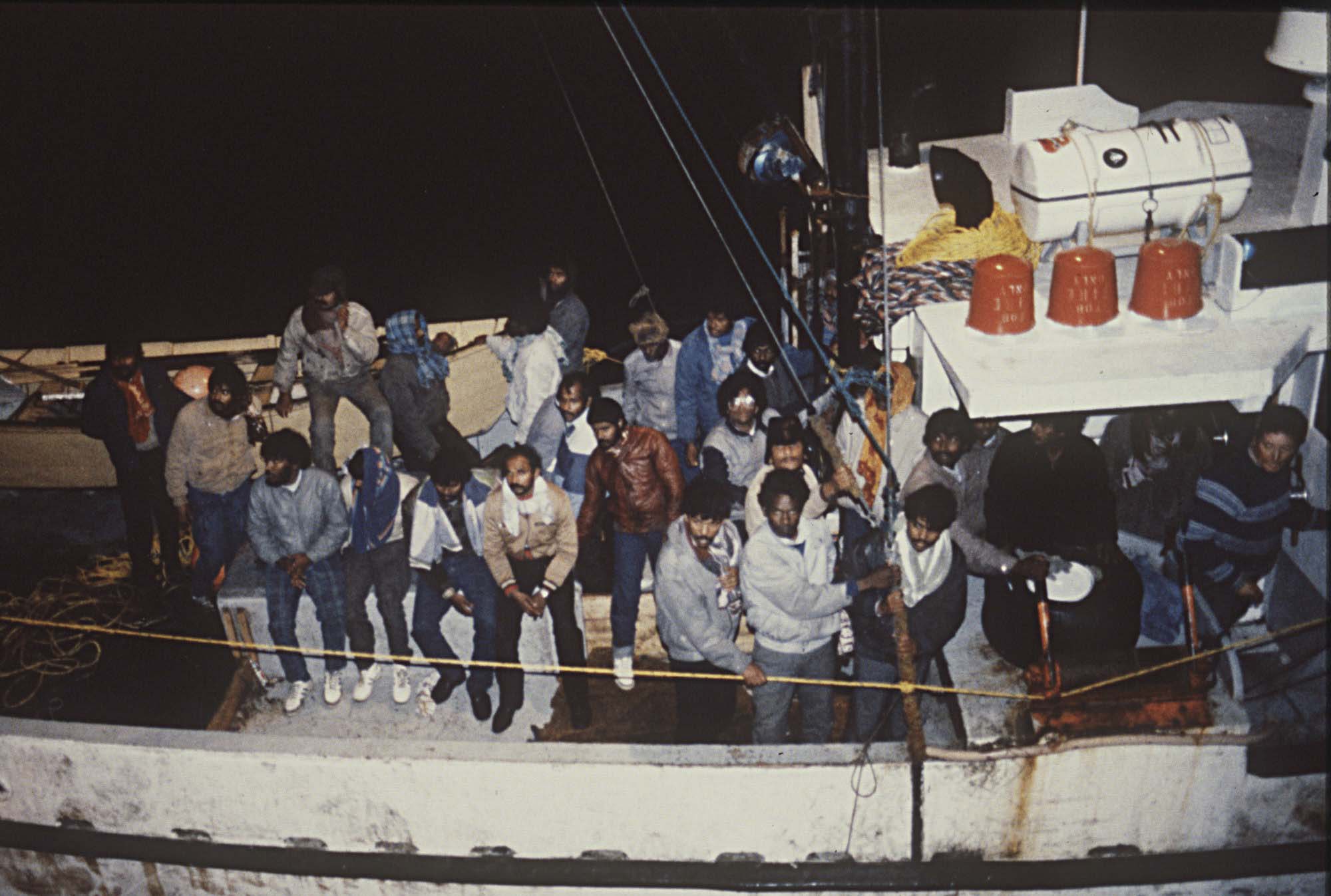 Un groupe d’hommes tamouls entassés à bord d’un petit bateau d’apparence ancienne. Le ciel que l’on peut voir derrière eux est complètement noir.