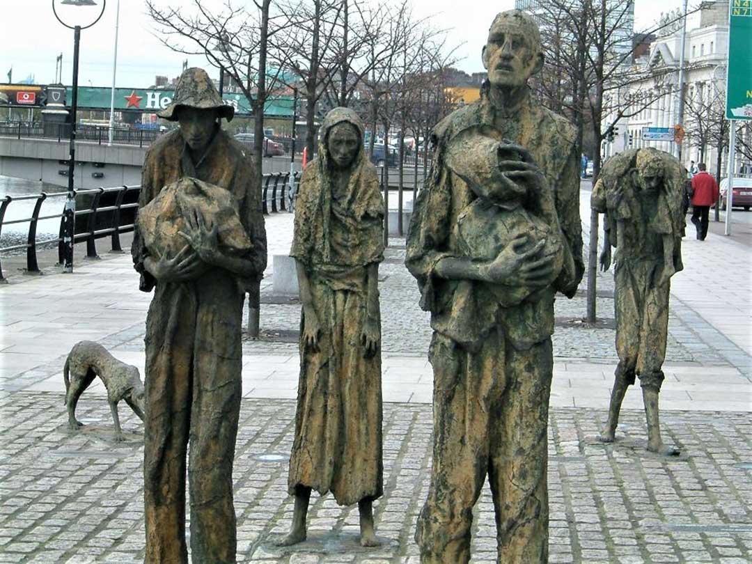 Des statues en bronze de marcheurs émaciés sur un large trottoir de la ville, un personnage porte un corps sur ses épaules.