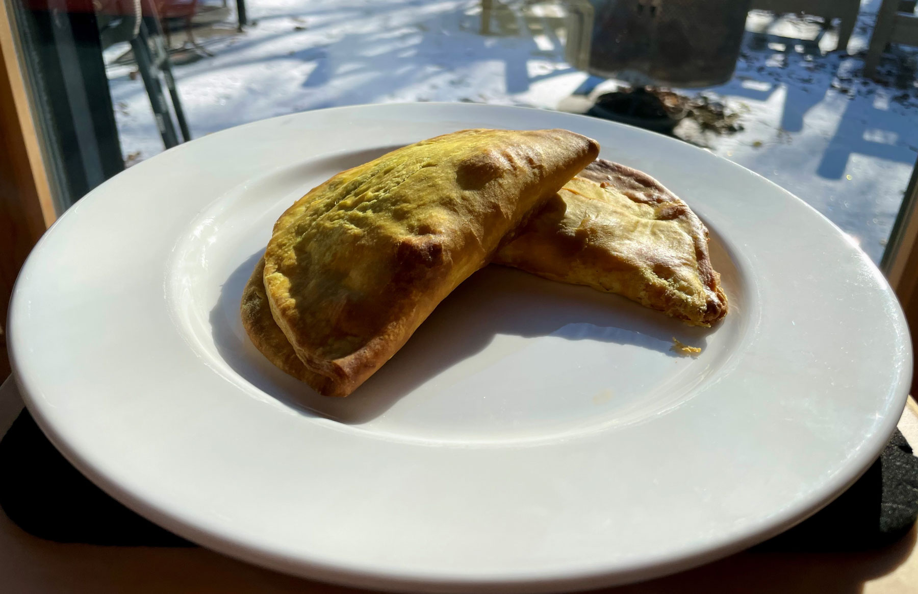 Des pâtés jamaïcains sur une assiette blanche, derrière l’assiette, en flou artistique, à travers une fenêtre, une fine couche de neige recouvre le sol.