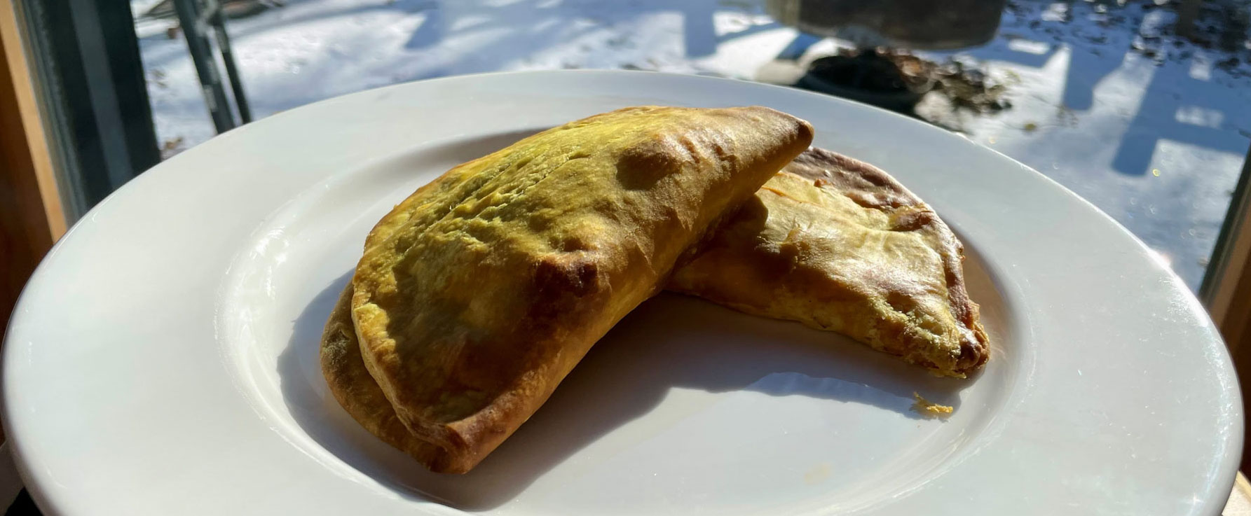 Des pâtés jamaïcains sur une assiette blanche. Derrière l’assiette, en flou artistique, à travers une fenêtre, une fine couche de neige recouvre le sol.