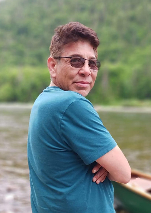Un homme au bord d’une rivière, près d’un canoë, regarde l’appareil photo par-dessus son épaule.