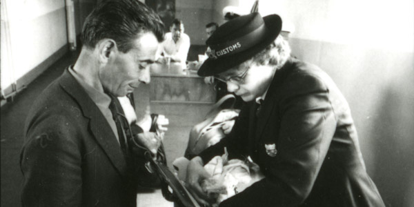 Dans une vieille photo en noir et blanc, un douanier fouille dans le sac d’un homme en complet.