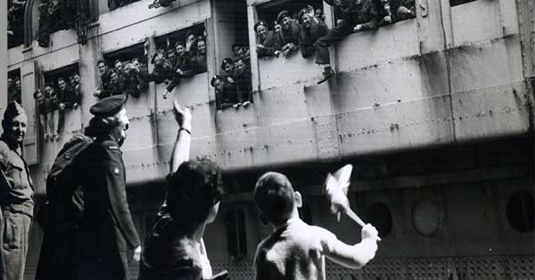 Ancienne photo en noir et blanc du temps de la guerre montrant un navire de transport de troupes accostant. Des centaines de soldats envoient la main et un petit garçon et sa mère leur envoient la main en retour à partir d’une plateforme. La photo montre aussi un militaire regardant la scène.