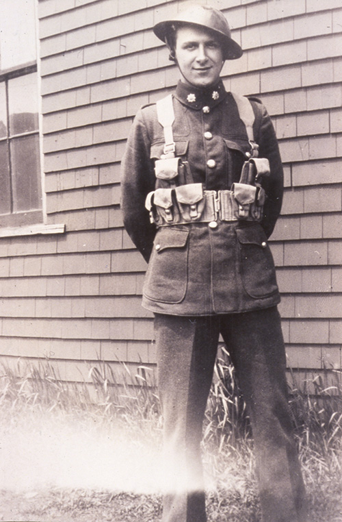 A man in a WWII Canadian army uniform including helmet stands proudly with his arms behind his back, the backdrop is the clapboard wall of a house