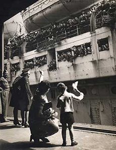 Une vieille photo en noir et blanc du temps de la guerre montrant un navire de transport de troupes amarré. Des centaines de soldats envoient la main et un petit garçon et sa mère leur envoient la main en retour à partir d'une plateforme. La photo montre aussi un militaire qui regarde la scène.