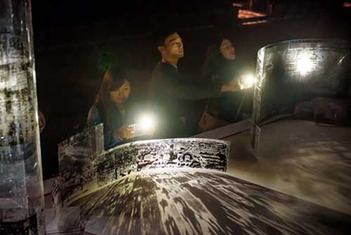 Trois personnes font briller les lumières de leurs téléphones à travers des formes transparentes courbées imprimées d’encre noire, créant des ombres sur la table.