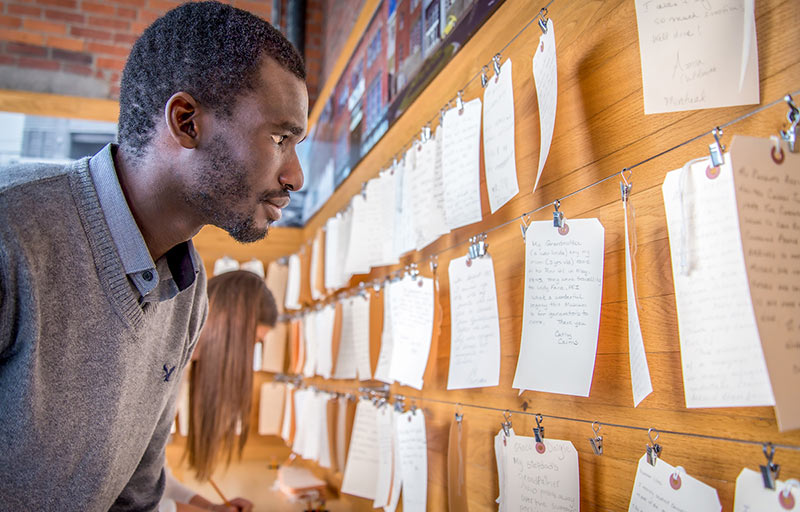 Un homme noir vêtu d'une chemise à col et d'un chandail lit des notes écrites à la main sur des étiquettes de bagages en papier accrochées à un mur.