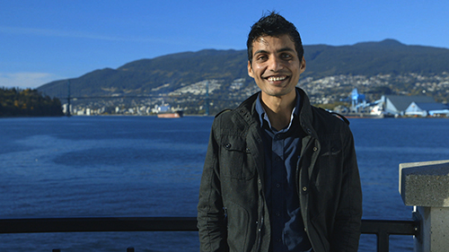 Un homme souriant debout contre une balustrade, derrière lui il y a de l'eau, un paysage urbain et une montagne.