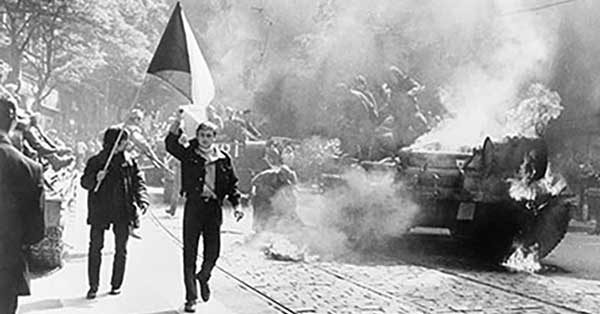 Une photo en noir et blanc de deux hommes sur un trottoir, l’un tenant un drapeau tchécoslovaque, alors qu’un char brûle dans la rue à côté d’eux.