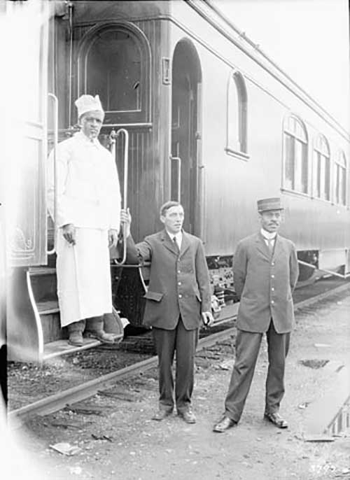 Photo noir et blanc de 3 hommes, un en uniforme de chef, un en costume et un en uniforme de porteur de train devant un wagon de train.