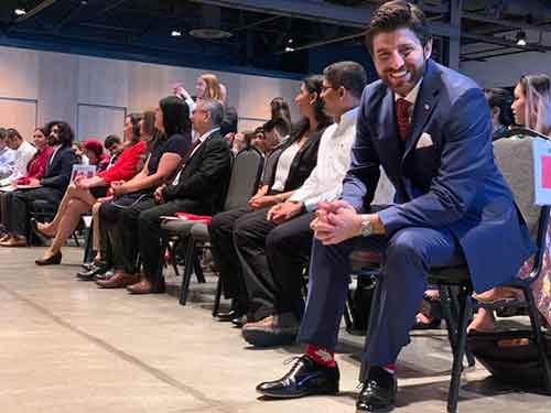 Un homme portant un costume bleu et une cravate rouge est assis dans une pièce avec beaucoup d’autres personnes, il sourit en direction de l’appareil photo.