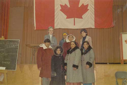 Un groupe de personnes, dont plusieurs portent des manteaux d'hiver doublés de fourrure, se tient devant un grand drapeau canadien.