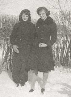 Une photographie en noir et blanc de deux jeunes femmes qui portent des manteaux d'hiver. Elles ont de la neige jusqu'aux chevilles et des arbres et des buissons sont en arrière-plan.