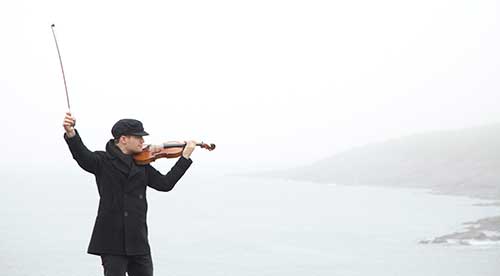 Un homme portant une veste et un chapeau de couleur sombre tient un violon et un archet. Un rivage océanique se trouve en arrière-plan.