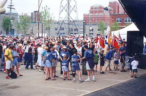 Une grande foule est rassemblée dans un stationnement. Les gens se trouvent près d’une ligne de pavillons, de haut-parleurs et d’une tente pour événement.