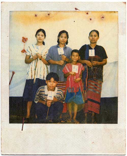 Une photo polaroid d'une famille de 5, mère, deux filles et deux fils, posent contre un fond de bâche blanche, chacun tenant un petit signe avec un nombre, 1 à 5.