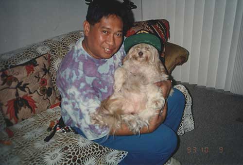 Un homme aux cheveux noirs vêtu d'une chemise tache-teinture berce un chien aux cheveux longs. Le chien porte une casquette de baseball.