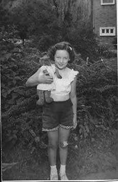 Une jeune fille en blouse blanche et en culottes courtes. Elle est dans un jardin et tient un ourson en peluche qui porte également une robe blanche.