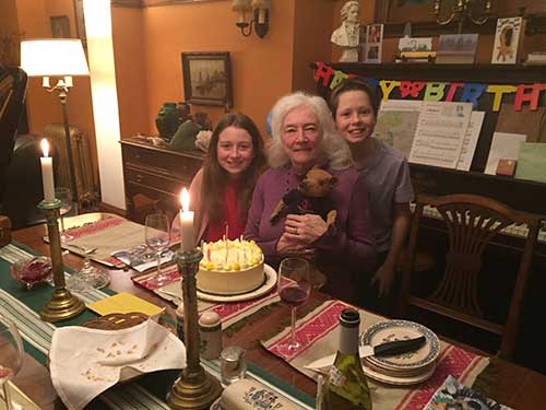 Une femme âgée assise tient un ourson en peluche. Une jeune fille et un jeune garçon sont à côté d’elle et devant elle se trouve un gâteau d’anniversaire jaune.