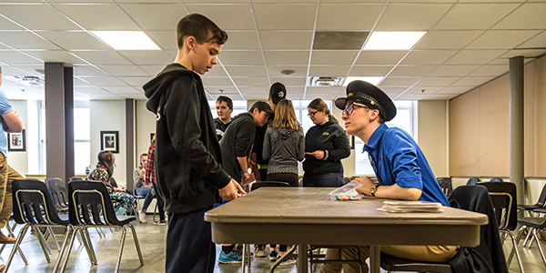 Un garçon est debout à une table. De l’autre côté de la table, un membre du personnel du Musée portant la casquette d’un douanier écoute attentivement le garçon.