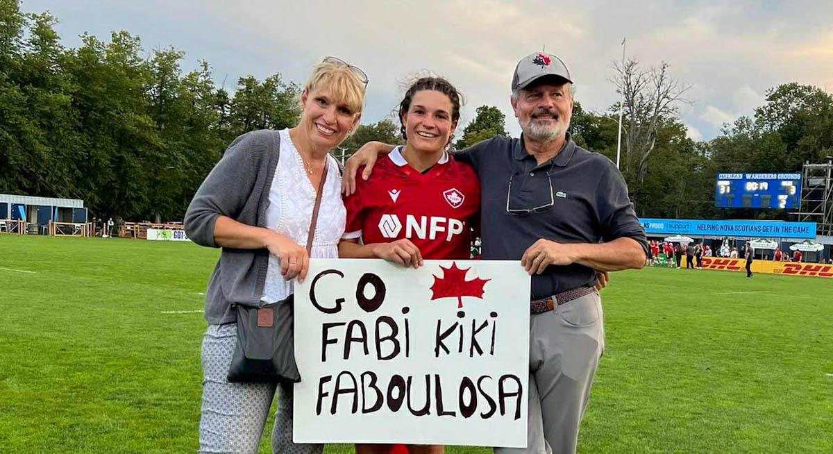 Un couple d’âge moyen se tient debout avec une jeune femme portant un uniforme de Rugby Canada, sur un terrain. Ils tiennent une pancarte sur laquelle on lit Go Fabi Kiki Fabulosa.