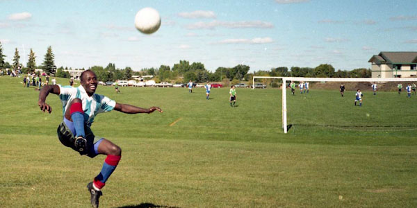 Un homme noir dont les pieds ne touchent plus le sol sourit après avoir botté un ballon. Il est en plein soleil, sur un grand terrain, devant terrain de soccer en activité.