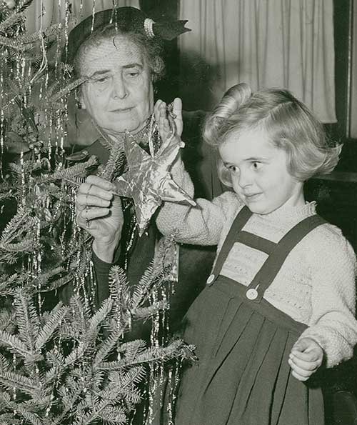 Une photo en noir et blanc d'une jeune fille suspendant un ornement à un sapin de Noël, une femme l'aide à placer l'ornement sur l’arbre.