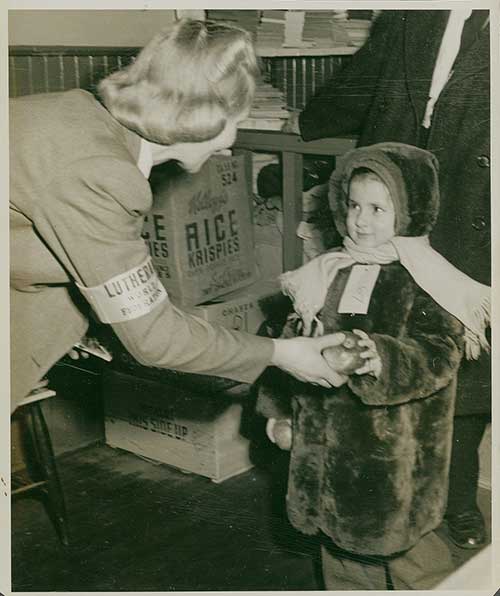 Photographie en noir et blanc d'une jeune fille vêtue d'un manteau d'hiver tenant une pomme offerte par une femme.