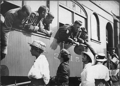 Vue latérale d'un train, des hommes en uniforme de la Première Guerre mondiale se penchent par les fenêtres pour dire au revoir aux femmes sur le quai