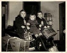 Image en noir et blanc sur laquelle se trouvent trois enfants assis avec des sacs de voyage. Ils se tiennent les uns les autres et semblent très heureux.