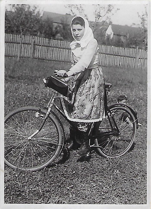 Photographie en noir et blanc d'une jeune fille sur un vélo. Elle est vêtue d'une jupe et d'un chandail à manches longues et porte un foulard sur la tête.