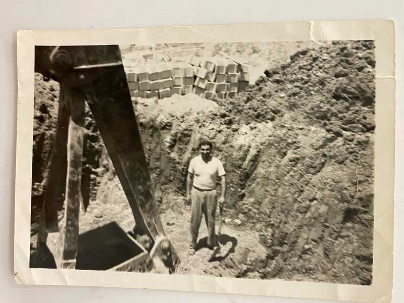 Young man stands in a pit, there are concrete blocks behind him and an excavator in front.