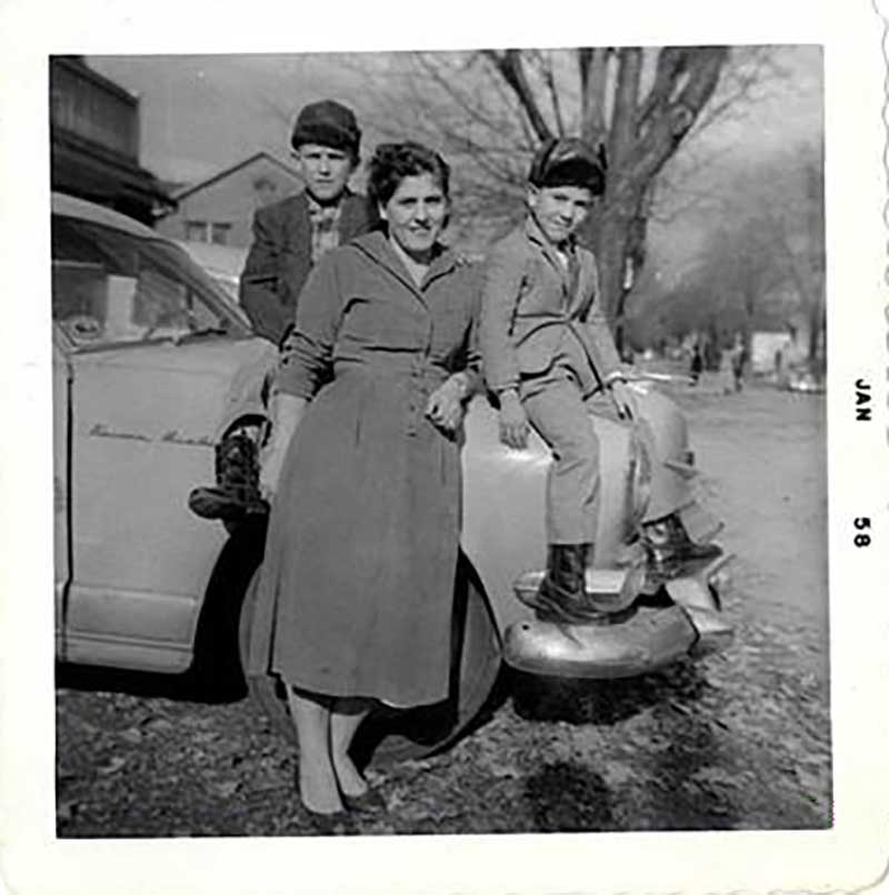 Une femme s’appuie contre une voiture avec deux garçons assis sur le capot.