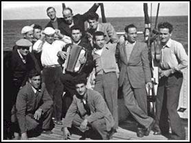 Bruno jouant de l’accordéon au milieu de plusieurs jeunes hommes sur le pont d’un bateau. 