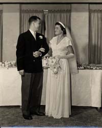 Photographie des époux le jour du mariage, devant une table de banquet. 
