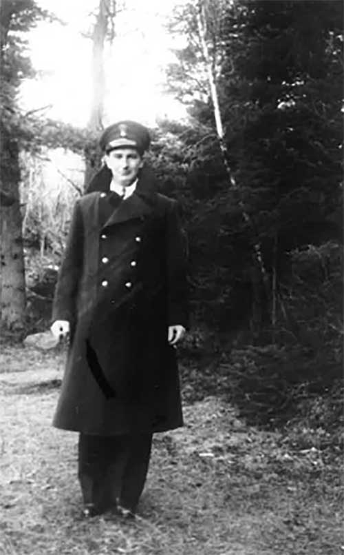 Un homme en uniforme devant un arbre. Il porte un long manteau noir et une casquette.