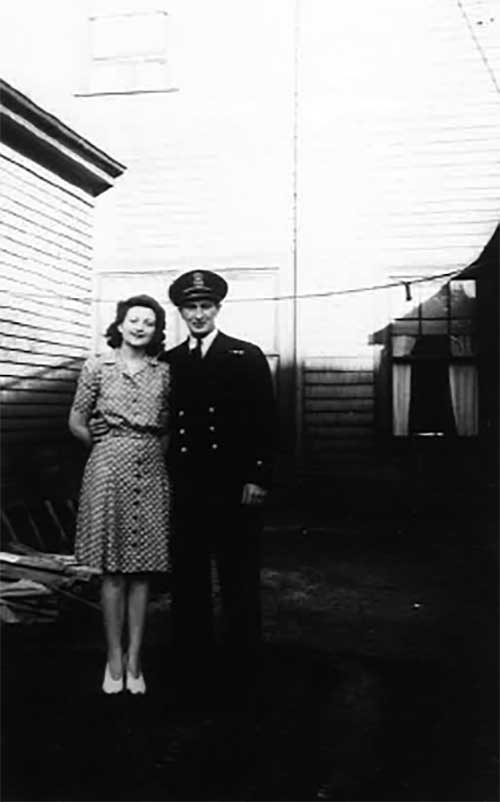 Un jeune couple devant une maison. La femme porte une belle robe et l’homme est en uniforme.