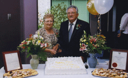 Pompilio et Rosa debout derrière une table garnie d’un gâteau, de fleurs et de ballons. 