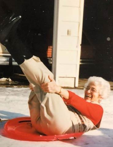 Une femme blanche âgée, dehors, sur une luge circulaire, riant et tenant ses jambes en l’air.
