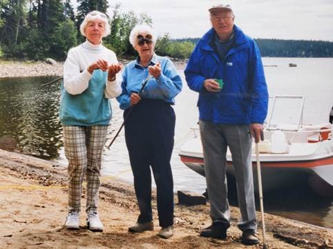 Trois personnes blanches d’un certain âge, deux femmes et un homme, au bord d’un lac, avec des bâtons et un bateau derrière eux.