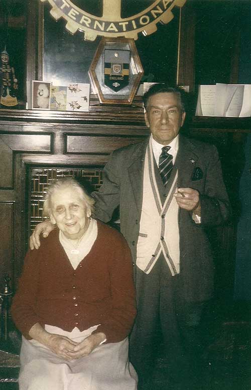 Une femme âgée est assise sur une chaise, un homme se tient à côté d’elle devant une cheminée.