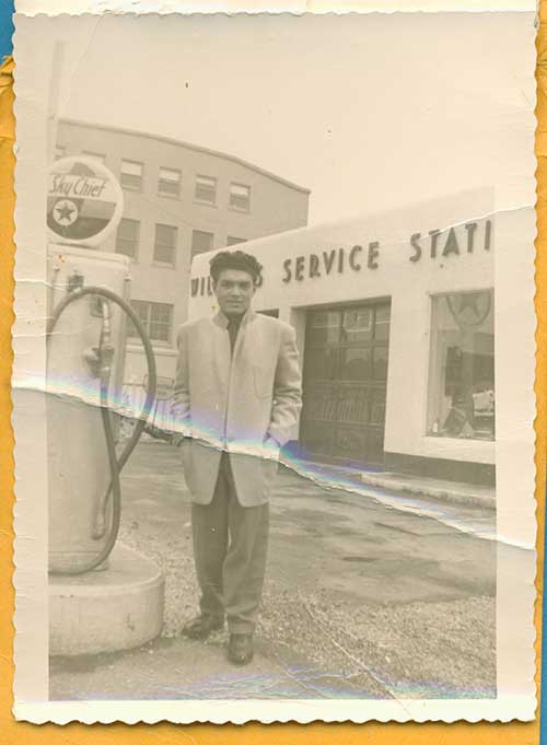 Un jeune homme se tient devant une station-service à côté d’une pompe à essence.