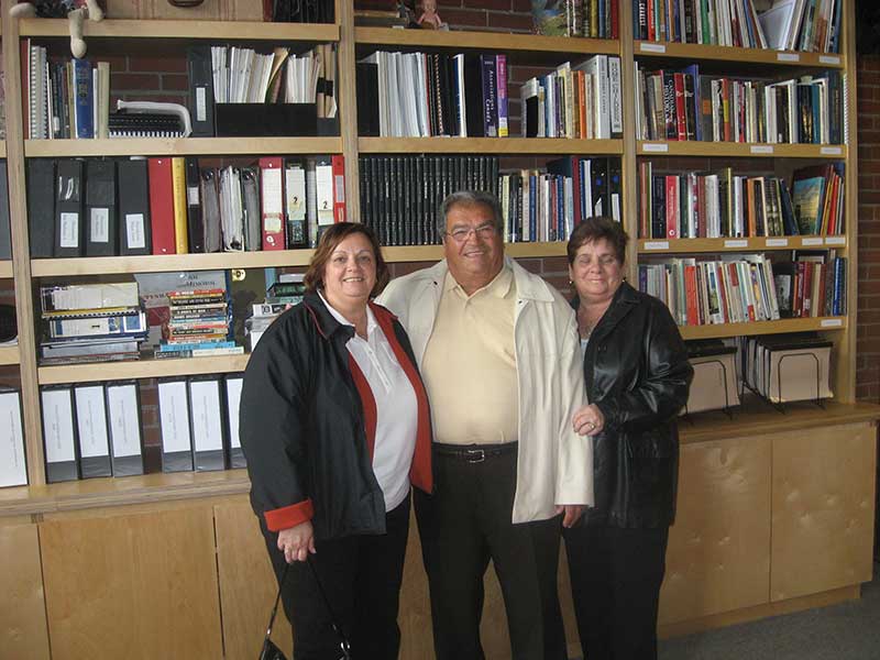 Un jeune homme avec deux femmes se tient à côté d’une grande étagère à livres.