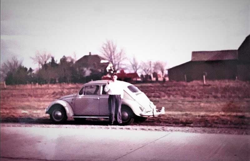 Un jeune homme appuyé contre une Beetle de Volkswagen blanche. Il regarde en direction de l’appareil photo qui est situé de l’autre côté de la rue.