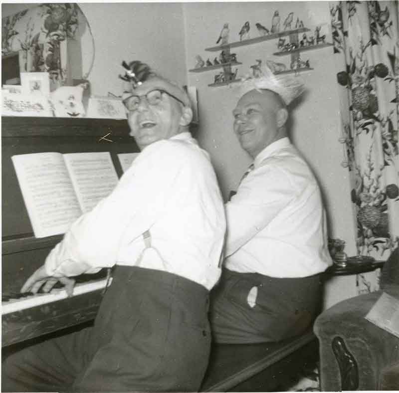 Deux gentlemen assis devant un piano. Ils rient et chantent.