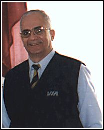 Homme debout avec des lunettes teintées et portant une veste bleue avec VIA sur la poitrine.