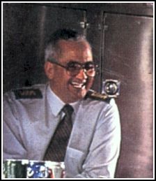 Un homme qui rit, qui porte des lunettes teintées, une chemise blanche et une cravate.
