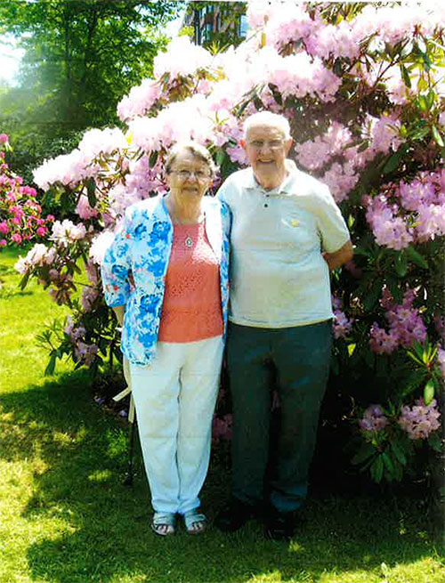 Un couple de personnes âgées se tient devant un magnifique buisson fleuri alors qu’ils posent pour une photo.