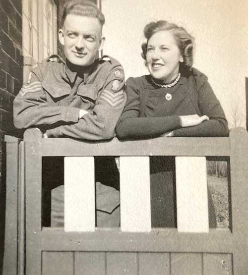 Photo en noir et blanc d’un homme en uniforme et d’une jeune femme bien habillée. Ils sont tous deux penchés sur la clôture en bois et debout avec les bras croisés.