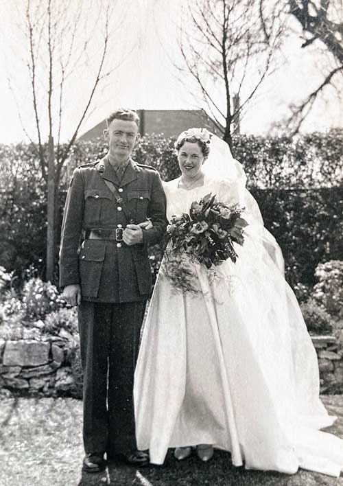 Une jeune mariée portant une robe de mariée et tenant des fleurs se tient à côté d’un jeune homme portant un uniforme.
